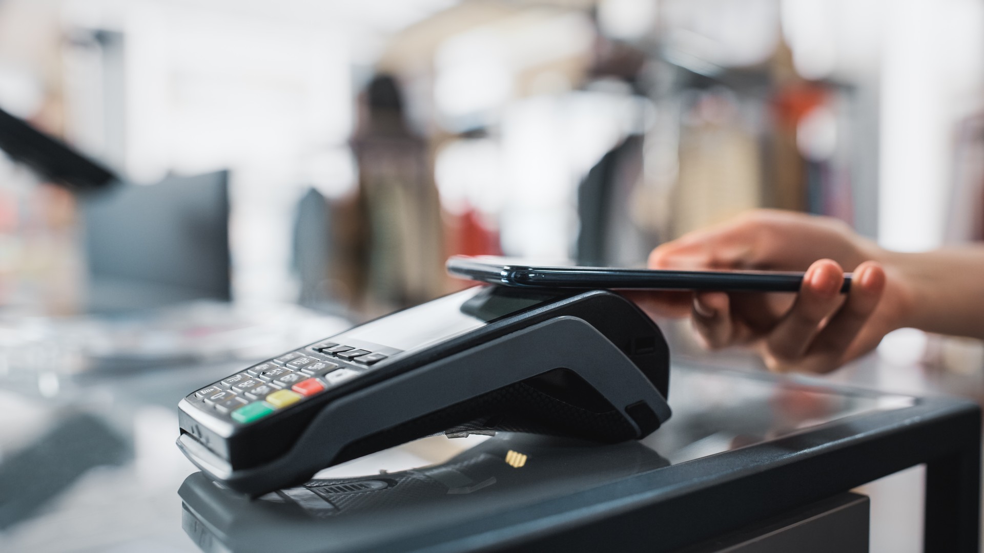 Clothing Store: Customer at Counter Buys Clothes Paying with Smartphone Through Contactless NFC Bank Terminal. Trendy Fashion Shop with Stylish Designer Brands. Close-up Focus on Hand.